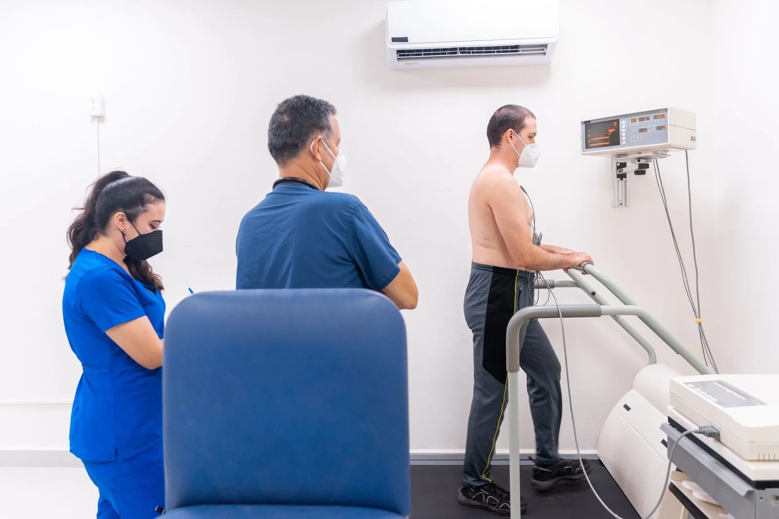 Man som går på löpband under ett konditionstest med EKG-elektroder, medan personal följer testet.