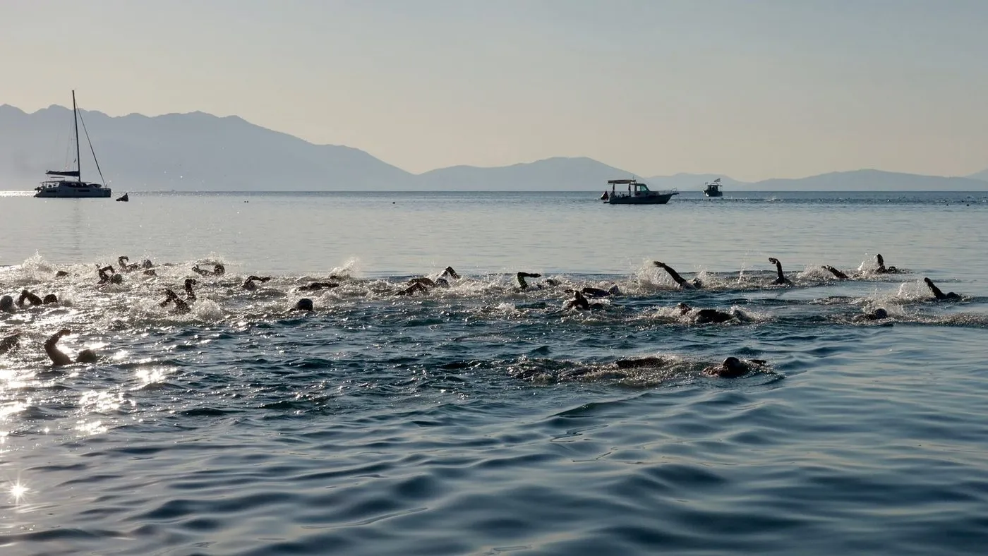 Triathleter simmar i öppet vatten i morgonljus med båtar och berg i bakgrunden