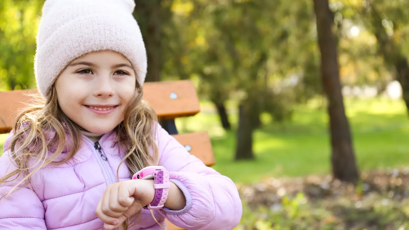 Liten flicka med rosa mössa och rosa barnklocka sitter på en parkbänk och tittar på handleden