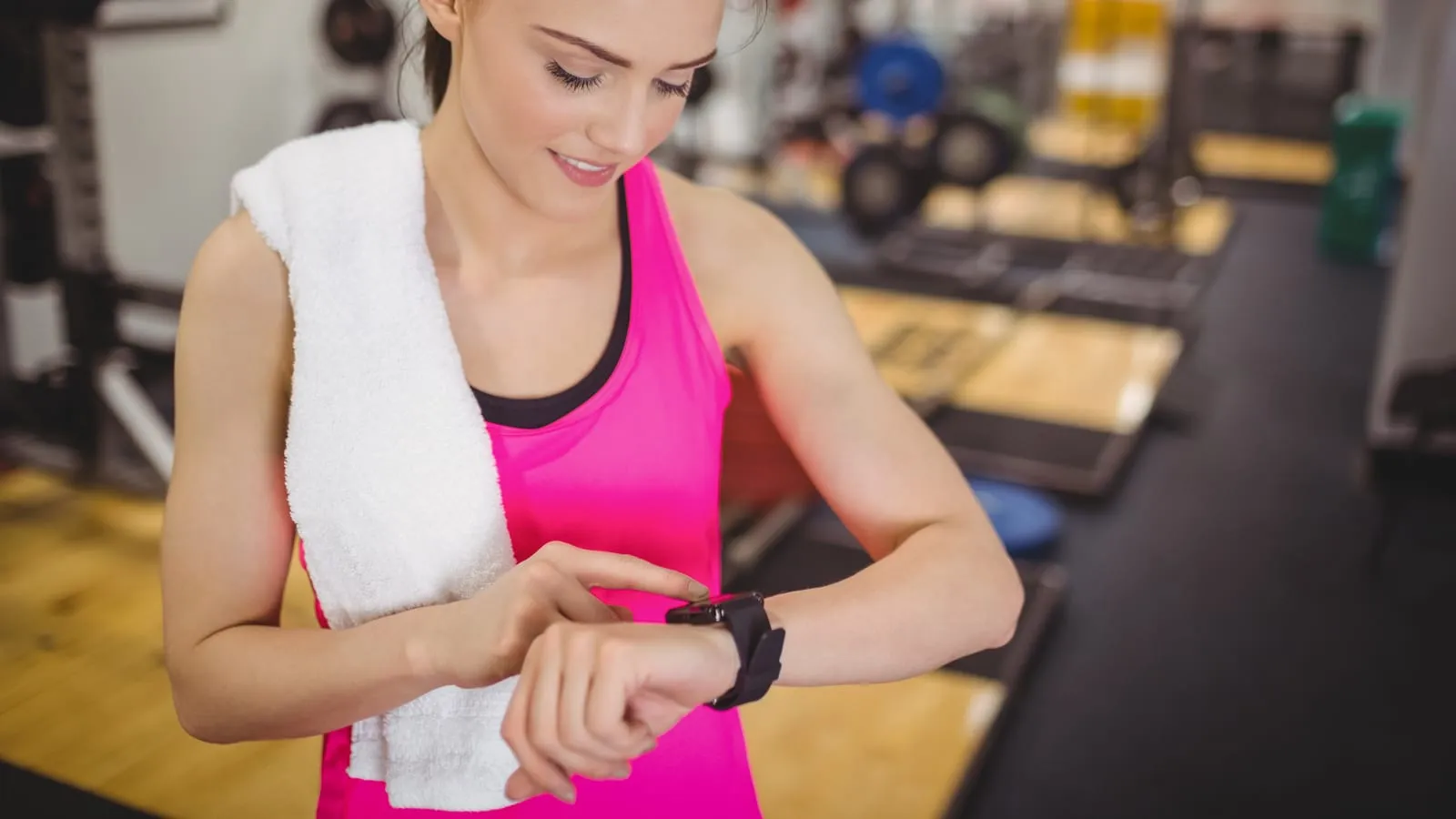 Kvinna med handduk runt halsen kontrollerar en sportklocka på handleden i ett gym